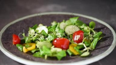 女人在蔬菜沙拉中加入盐与蔬菜，黄瓜，西红柿，胡椒。 把落在石盘上的绿色沙拉混合在一起